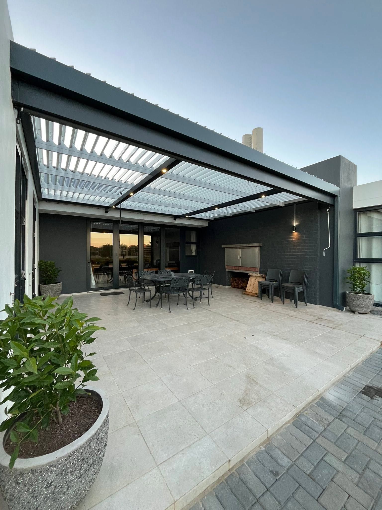 Charcoal louvre awning with white louvres over a paved entertainment area at dusk